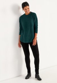 Türkiser Strickpullover mit rundem Ausschnitt, langen Ärmeln und leicht geschwungener Saumlinie, kombiniert mit schwarzen Slim-Fit-Hosen und Knöchelstiefeln.