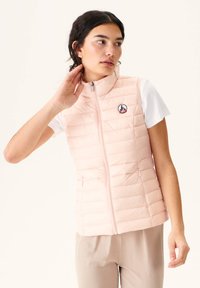 Light pink puffer vest with horizontal quilted design, front zipper, and round logo patch. Worn over a white T-shirt, paired with light pants.