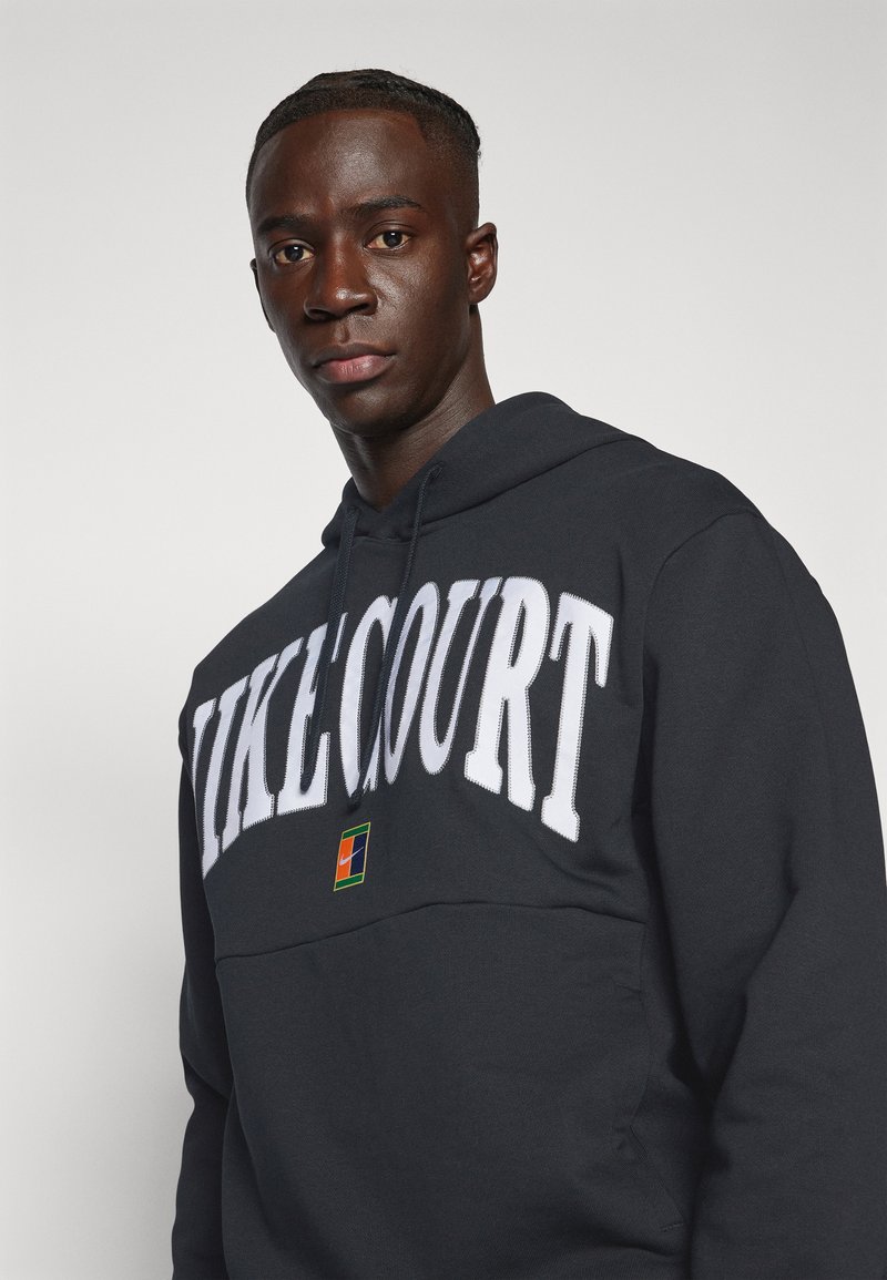 Felpa nera con la scritta bianca "NIKE COURT". Presenta una tasca a marsupio e un piccolo logo colorato sul petto. Texture morbida, vestibilità casual.