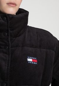 Černá kordurová bunda s vysokým límcem, vybavená černými knoflíky a obdélníkovým logem Tommy Jeans na hrudi.