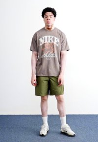 Brun grafisk T-shirt med et Nike-logo, grønne shorts, hvide strømper og grå sneakers. Enkel design, neutrale farver, afslappet stil.