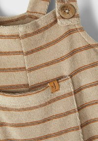 Beige Baumwolloveralls mit braunen und grauen Streifen, ausgestattet mit einer Brusttasche und einem Holzknopf-Detail am Träger.