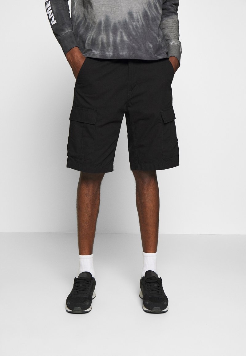 Homme portant un short cargo noir, des chaussettes blanches et des baskets noires, les mains dans les poches et un t-shirt gris à manches longues.