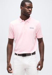 Homme portant un polo Calvin Klein rose clair, pantalon blanc et ceinture sombre, regardant vers sa gauche sur un fond clair uni.