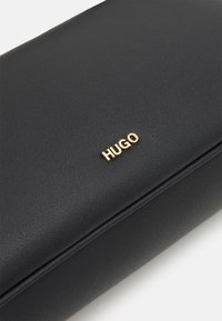 Bolso de mano negro con textura y un acabado suave, que presenta un prominente logo dorado "HUGO" en la parte frontal. Diseño rectangular simple.