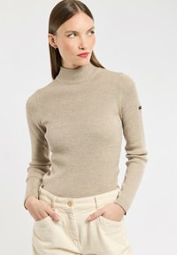 Femme aux longs cheveux bruns portant un sweater beige à col roulé côtelé et un pantalon crème taille haute, les mains dans les poches, regardant sur le côté devant un fond blanc.