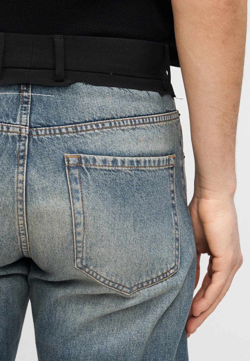 Gros plan d'une personne portant un jean en denim bleu délavé avec une poche arrière et une ceinture noire sur une chemise noire.