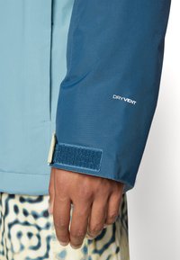 Rukáv vodoodolnej bundy v tmavomodrom materiáli, s uzáverom na suchý zips a logom "DRY VENT" v bielej farbe. Hladká textúra a športový strih.