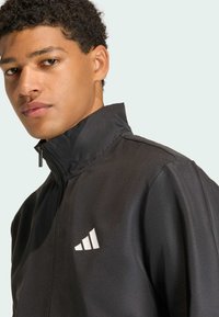 Μαύρο τζάκετ με φερμουάρ και ψηλό γιακά, κατασκευασμένο από λείο ύφασμα; διαθέτει λευκό λογότυπο στην αριστερή περιοχή του στήθους; μινιμαλιστικός σχεδιασμός.