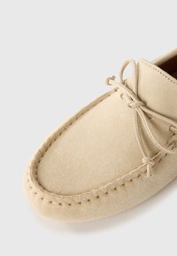 Mocassin en suède beige avec un bout rond, des bords cousus et des lacets décoratifs. Texture douce et design minimaliste.