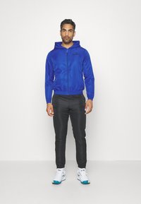 Kék cipzáras dzseki kapucnival, rugalmas alsó szeg�éllyel és húzózsinórral. Fekete, slim-fit nadrág. Fehér cipők türküszínű akcentusokkal. Egyszerű háttér.