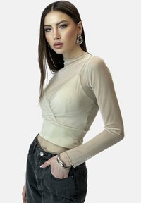 Beige omlottopp med långa ärmar i genomskinligt tyg, figurnära form och rundad fåll, kombinerad med mörka jeans med hög midja; accessoarer inkluderar silverörhängen.