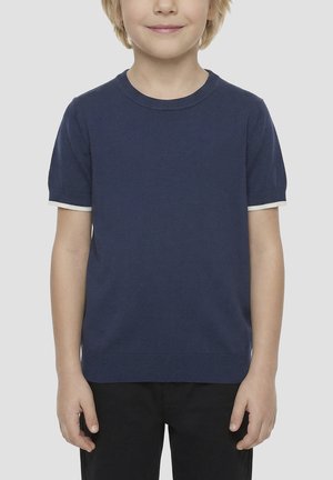 Malo dijete se smiješi, nosi mornarskoplavu majicu kratkih rukava s bijelim rubom na rukavima i crne hlače, stoji ispred jednobojne pozadine.