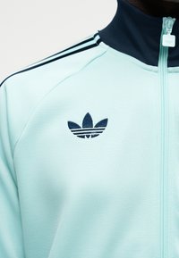 Svetlo modra Adidas trenirka z mornarsko modrim ovratnikom, tremi črtami na ramenih in vezenim logotipom trefoil na levem prsnem delu.