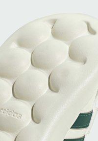 Suela de sneaker texturizada blanca con bultos redondeados, con la marca "adidas" visible. Parte superior de cuero verde y blanco con acentos a rayas.