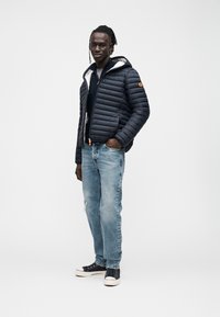 Tengerészkék steppelt bélelt dzseki kapucnival, szürke belső bélés, világoskék kopott farmerrel és fekete sportcipővel párosítva.