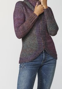 Femme portant un pull zippé en tricot violet et gris avec des bords côtelés et un jean bleu avec broderie florale sur la cuisse.