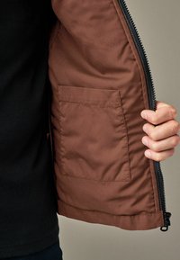 Personne tenant une veste marron ouverte, montrant une poche intérieure carrée et une fermeture éclair noire sur un fond neutre.