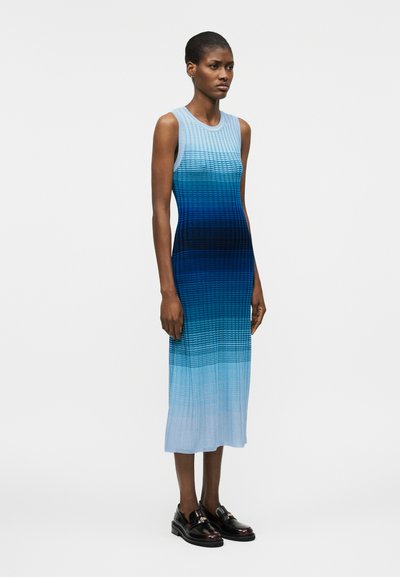 Λεπτή γυναίκα στέκεται φορώντας ένα μαν sleeveless, ριπώτο midi φόρεμα σε μπλε απόχρωση με διαβάθμιση και μαύρα loafers, μπροστά σε ένα απλό φωτεινό φόντο.