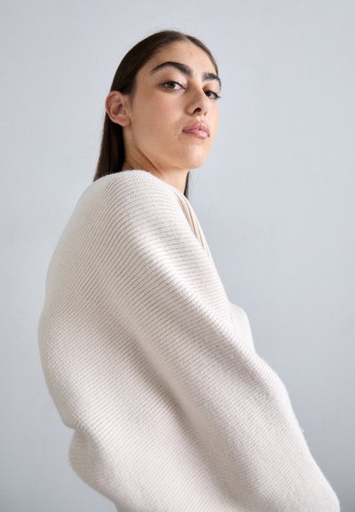 Gaiši bēšs, ribots adījums, kas pārlikts pār pleciem, ar mīkstu tekstūru un apjomīgu formu, papildināts ar minimālistisku fonu.