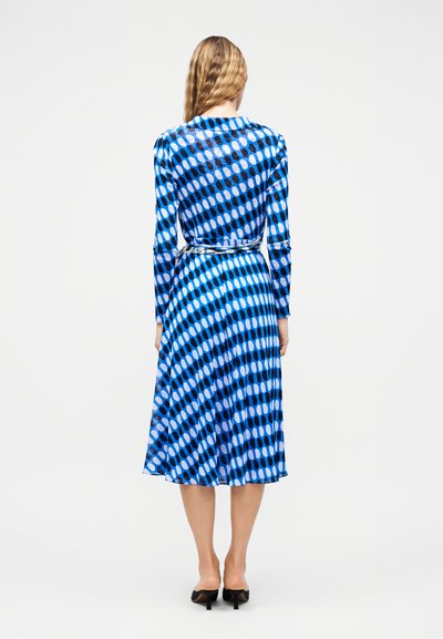 Γυναίκα με κυματιστά ξανθά μαλλιά, που φοράει μπλε midi φόρεμα με μακριά μανίκια και μοτίβο, και μαύρες γόβες, στέκεται γυρισμένη προς τα πίσω σε λευκό φόντο.