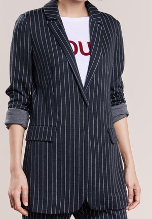 Vrouw die een marineblauwe krijtstreepblazer draagt met opgerolde mouwen over een wit overhemd met rode letters op een effen achtergrond.