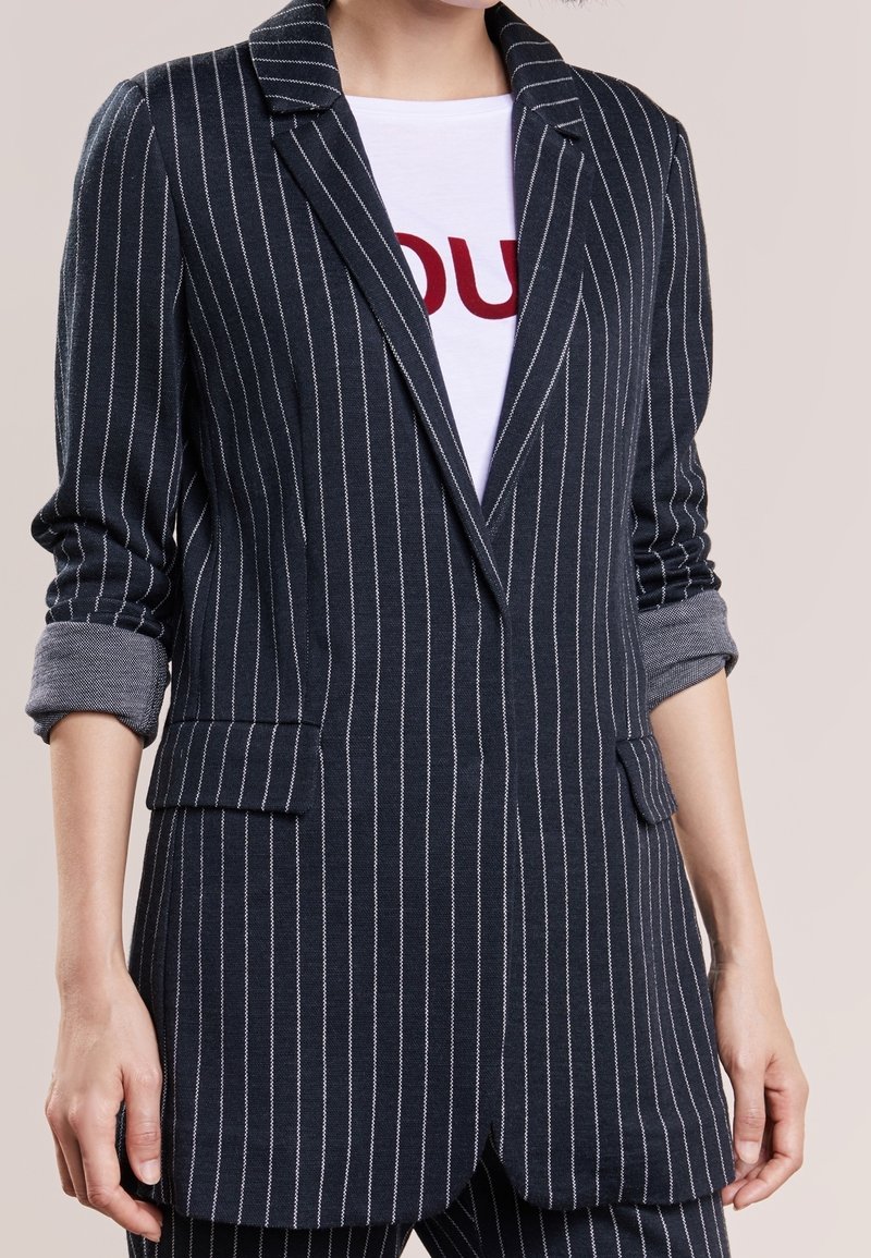 Femme portant un blazer bleu marine à fines rayures avec manches retroussées sur une chemise blanche avec des lettres rouges sur un fond uni.