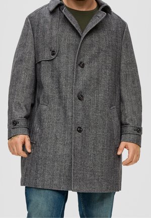 Homme portant un manteau gris à chevrons mi-long avec des boutons, une poche à rabat et des poignets, par-dessus une chemise verte et un jean bleu.