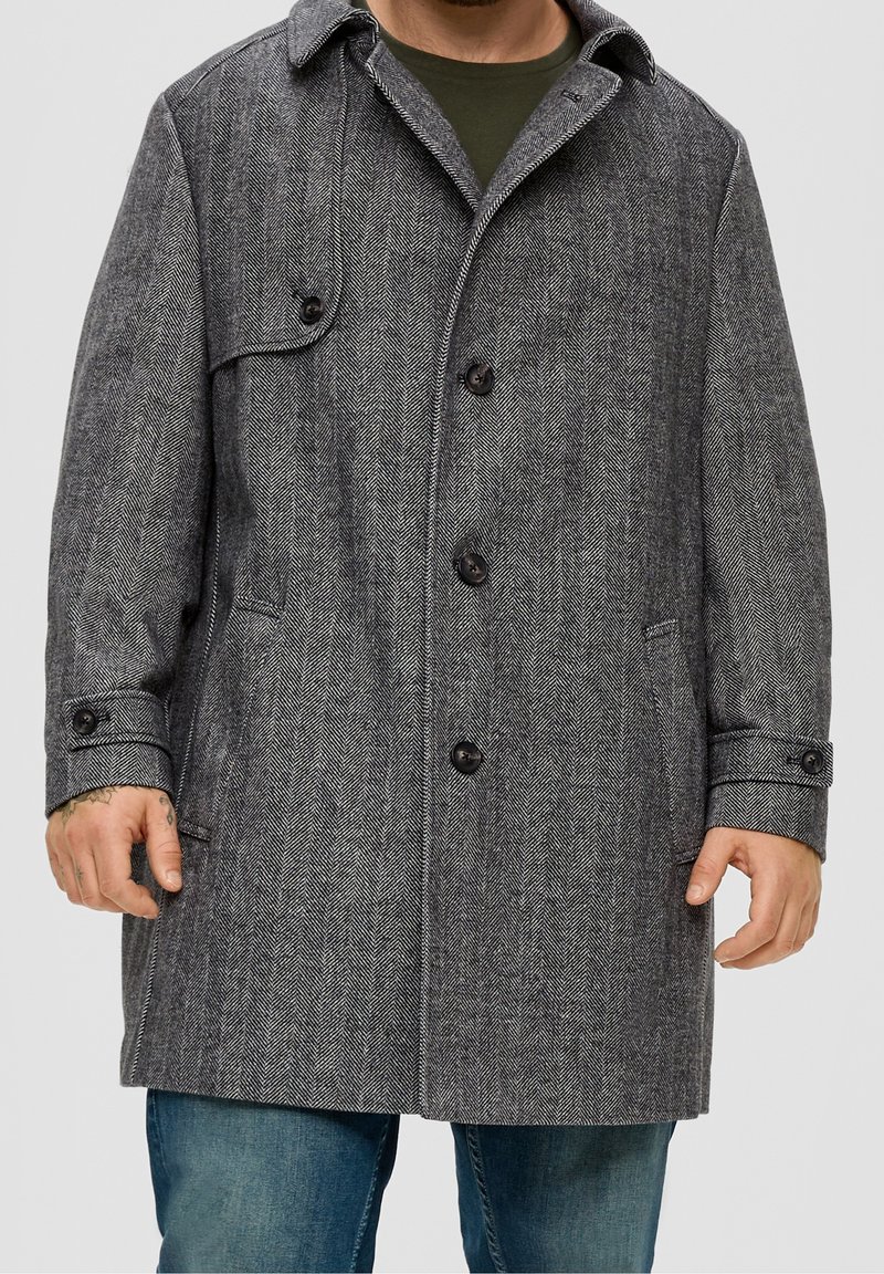 Homme portant un manteau gris à chevrons mi-long avec des boutons, une poche à rabat et des poignets, par-dessus une chemise verte et un jean bleu.