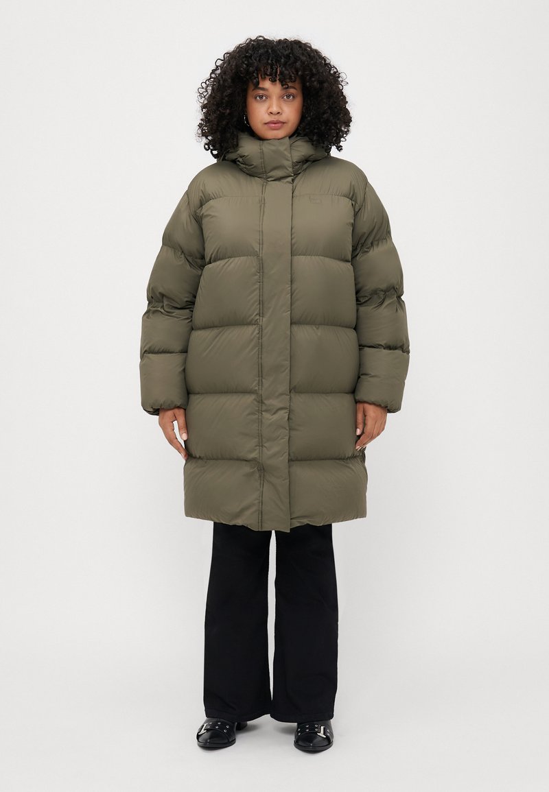 Geacă puffer verde măsliniu cu guler înalt, design structurat, segmente matlasate și mâneci lungi, purtată cu pantaloni negri cu croială largă.