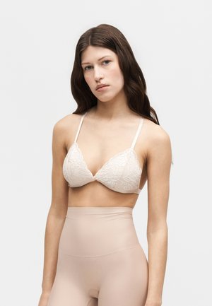 En ung kvinde med langt brunt hår har en hvid blonde-trekants-bh og højtaljede beige sømløse leggings på mod en ensartet lys baggrund.