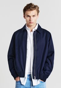 Giovane uomo che indossa una giacca zip navy sopra una camicia bianca con bottoni e jeans azzurri, in piedi con le mani in tasca su uno sfondo semplice.