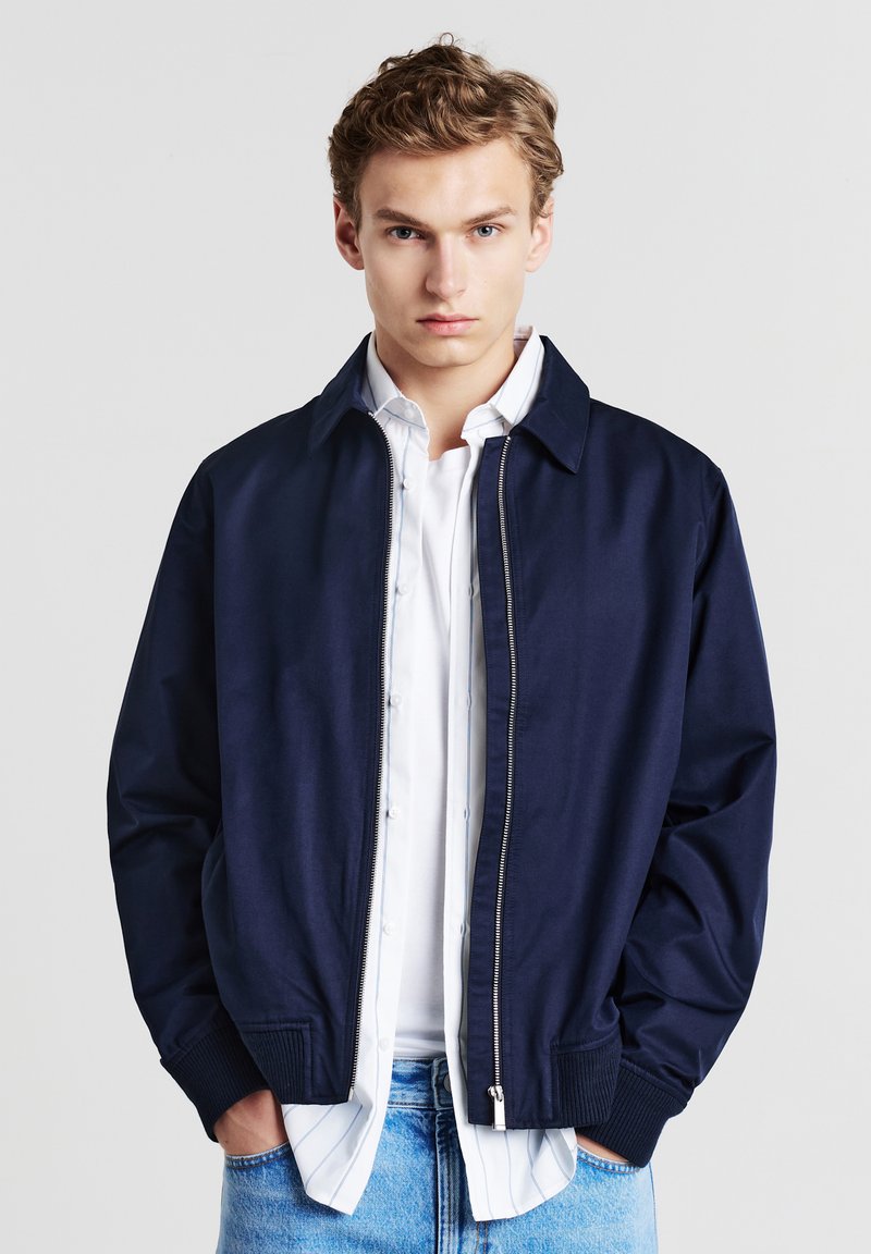Giovane uomo che indossa una giacca zip navy sopra una camicia bianca con bottoni e jeans azzurri, in piedi con le mani in tasca su uno sfondo semplice.