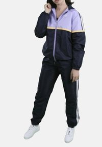 Tuta sportivo in viola e blu navy, con giacca zip-up e striscia gialla, pantaloni coordinati con strisce laterali e scarpe da ginnastica bianche.
