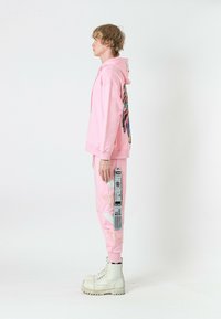 Sudadera rosa con un diseño de alas coloridas en la parte trasera, combinada con pantalones de chándal a juego que presentan detalles decorativos y una etiqueta prominente. Botas blancas.