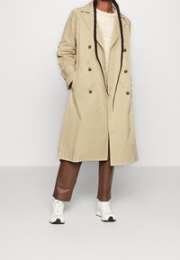 Beige double-breasted trenchcoat met grote bruine knopen, gedragen over een licht gestructureerde trui en bruine broek, gecombineerd met witte sneakers.