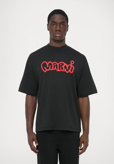 Hombre vistiendo una camiseta negra de gran tamaño con mangas cortas y un logo rojo de "Marni" en el pecho, de pie contra un fondo blanco liso.