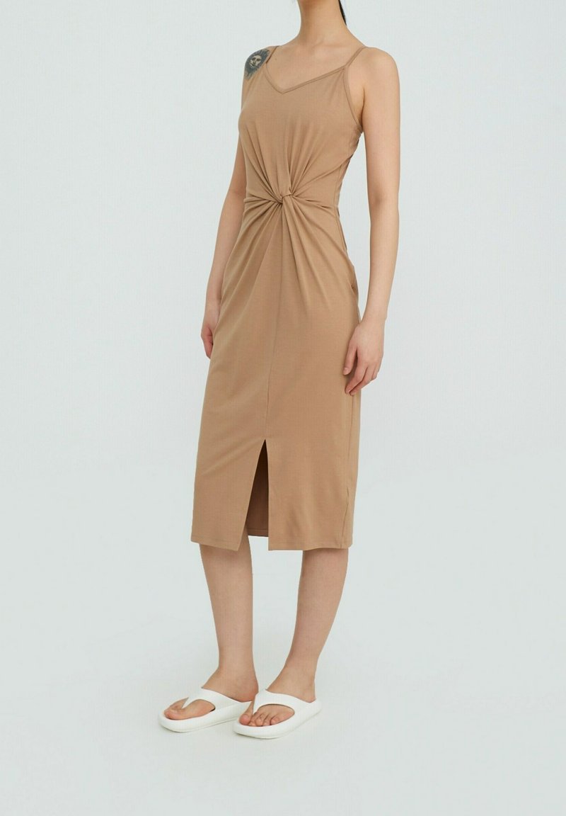 Robe midi beige ajustée avec un détail torsadé à l'avant, des bretelles fines réglables et une fente frontale, associée à des sandales tongs blanches.