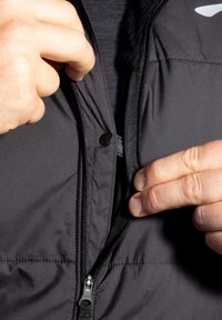 Schwarze gepolsterte Jacke mit einem Reißverschluss. Verfügt über einen Druckknopf am oberen Ende und ein weiches Innenfutter. Strukturierten Stoff ist sichtbar.