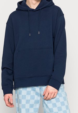 Navy blue hoodie with a front pocket, drawstring hood, and ribbed cuffs. Soft fabric, relaxed fit, and minimal design. Paired with checkered pants.