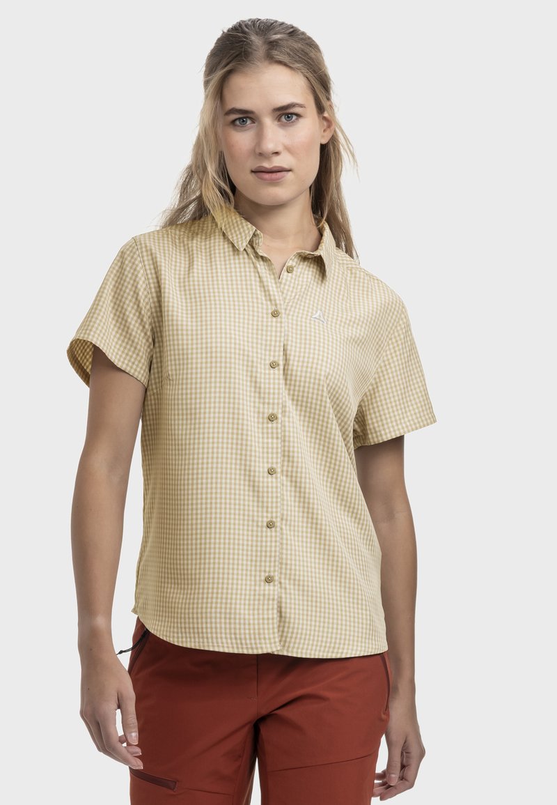 Young woman wearing short-sleeve beige checkered button-up shirt and rust-colored pants, standing against white background.