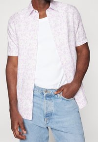 Koszula na krótki rękaw z guzikami w jasnofioletowym kolorze z kwiatowym wzorem, wykonana z lekkiego materiału. Nosi się ją na białym topie na ramiączkach, w zestawie z dżinsami.