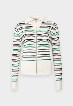 Kreminė zip-up megztinis su juodomis ir žaliomis horizontaliomis juostomis, apykakle, raukšlėtais rankogaliais ir apačia, ir mažas išsiuvinėtas logotipas ant krūtinės ir rankovės.