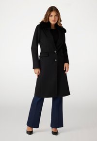 Cappotto di lana nero con colletto in pelliccia sintetica, due tasche frontali e bottoni argentati, abbinato a jeans blu a zampa d'elefante e tacchi neri.