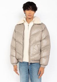 Man in een beige pufferjack met een witte schapenwollen kraag, een crèmekleurige gebreide ritstrui en blauwe spijkerbroek, staand tegen een effen witte achtergrond.