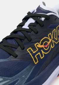 Μπλε αθλητικό παπούτσι με υφασμάτινο επάνω μέρος με υφή, μαύρα κορδόνια και έντονο κίτρινο λογότυπο "HOKA" με κόκκινη λεπτομέρεια. Είναι ορατό το εξωτερικό πέλμα με σχεδιασμό καπακιού.