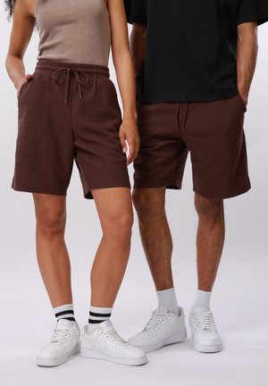 Dos personas con shorts de punto marrón a juego, zapatillas blancas y calcetines deportivos, de pie una al lado de la otra con las manos en los bolsillos.