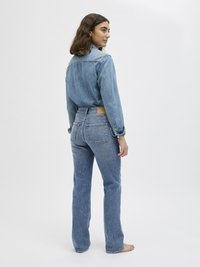 Zils denim krekls ar pogām priekšā, savienots ar gaiši ziliem platām bikšu. Redzamie šuvumi un kabatas; modelis stāv basām kājām, vērsts sānis.