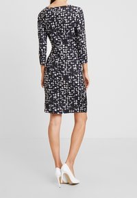 Robe noire avec un motif texturé de cercles blancs et gris, manches trois-quarts et longueur genou ; associée à des talons hauts blancs.