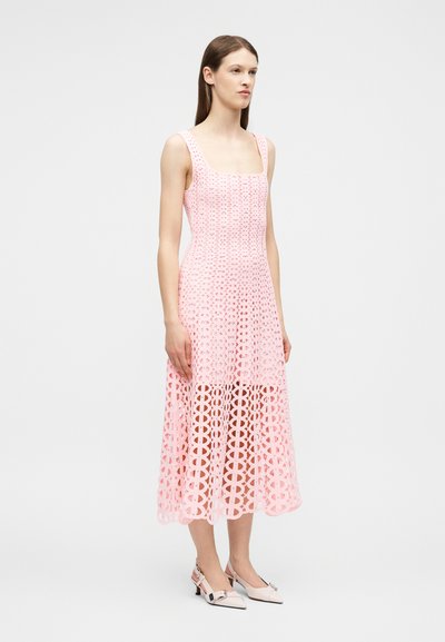 Femme portant une robe midi rose sans manches avec un motif de découpes circulaires et des escarpins slingback rose pâle, debout devant un fond uni.